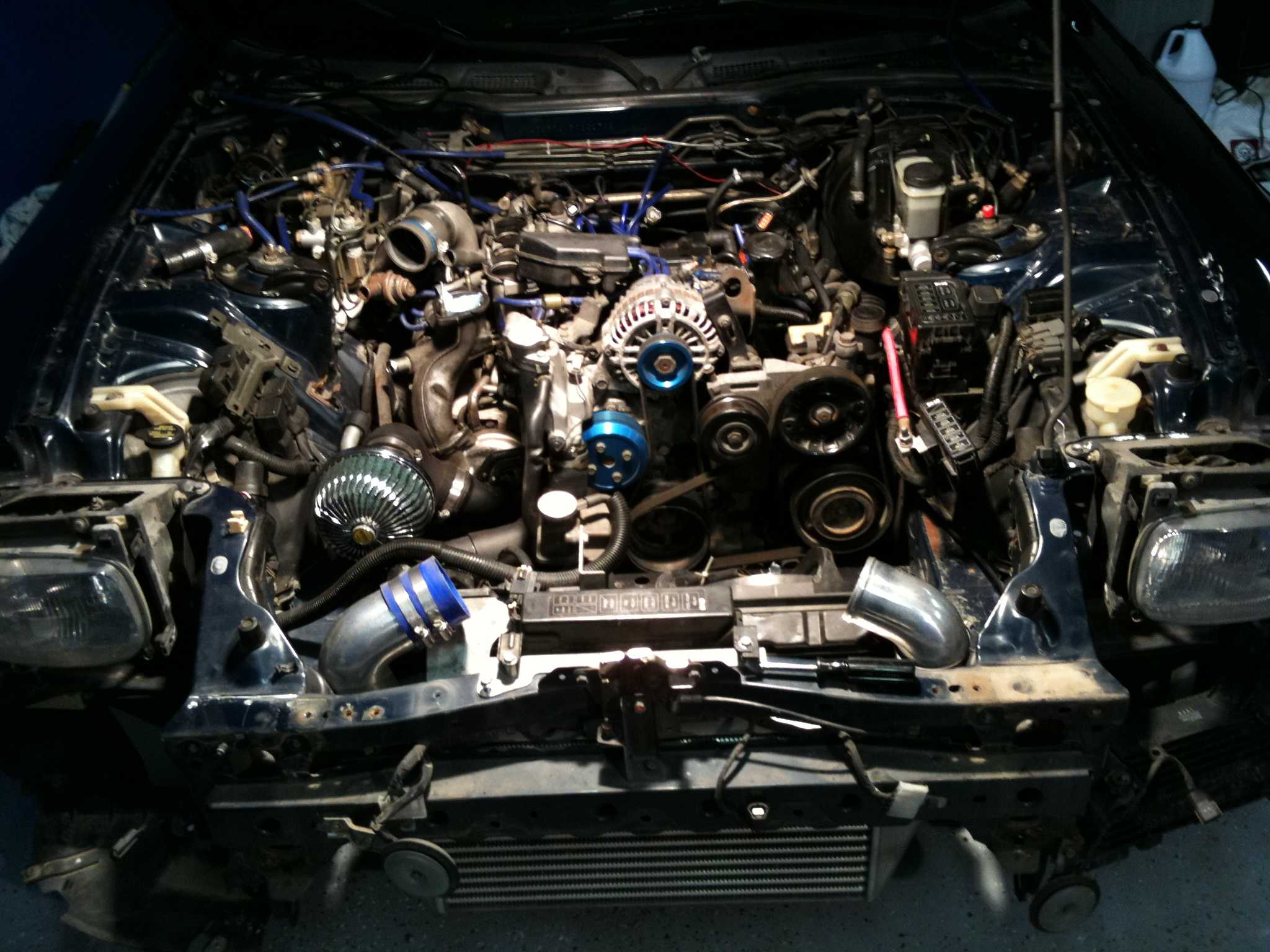 Bold charcoal drawing of a disassembled car engine bay showing wiring, hoses, and a turbo intake system, dark tones with blue accents