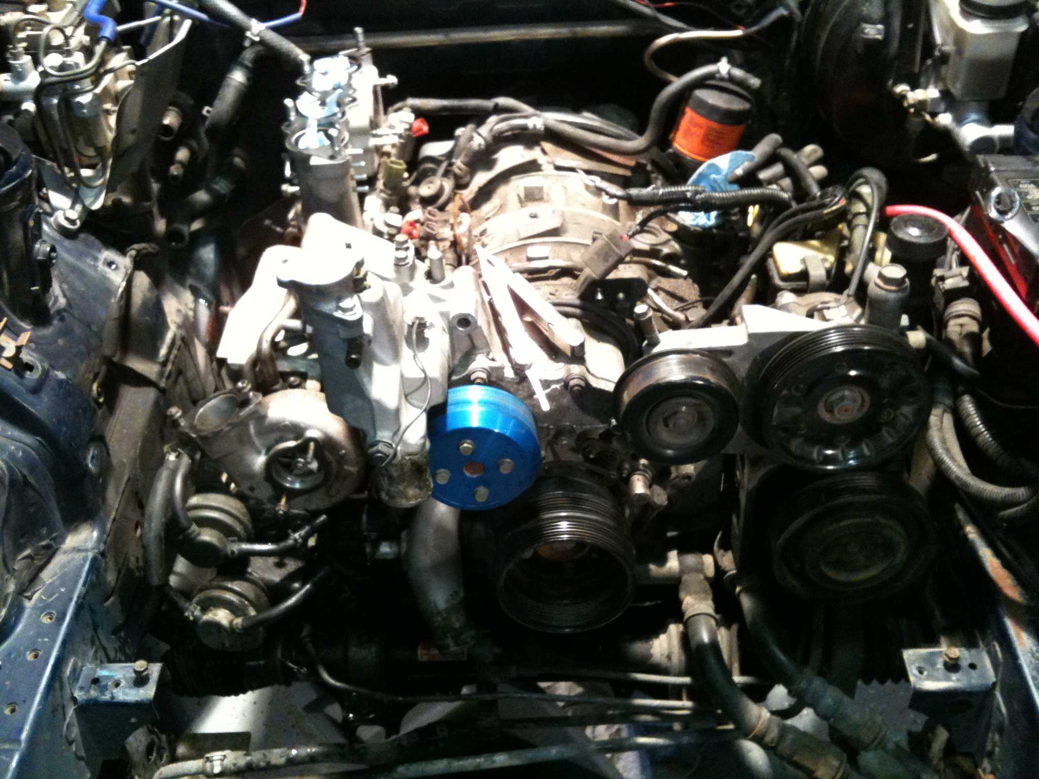 Wide view of an engine bay under repair with pulleys, belts, and a blue anodized pulley visible